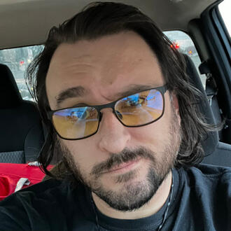 Selfie of a man with a short-trimmed beard, shaggy shoulder-length brown hair, and glasses, wearing a simple black t-shirt, sitting in a truck.